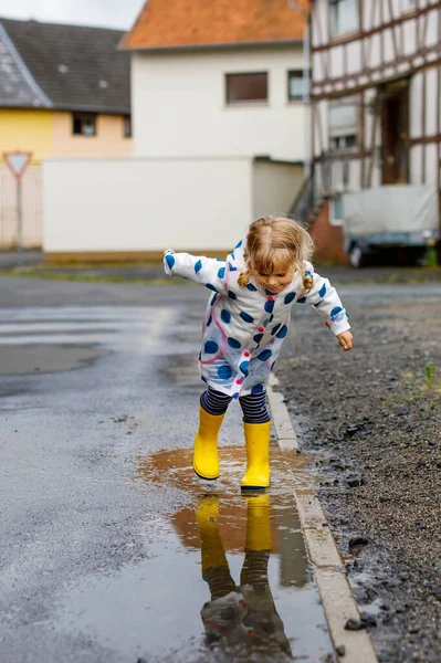 Malá batolata ve žlutých holínkách do deště, běhající a chodící během deštivého oblačného dne. Roztomilé šťastné dítě v barevných šatech skákání do kaluže, cákání vodou, outdoorové aktivity — Stock fotografie