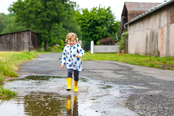 Kleines Mädchen in gelben Regenstiefeln, läuft und geht bei Schneeregen an einem regnerischen, bewölkten Tag. Nettes glückliches Kind in bunten Kleidern, das in eine Pfütze springt, mit Wasser planscht, Outdoor-Aktivität — Stockfoto