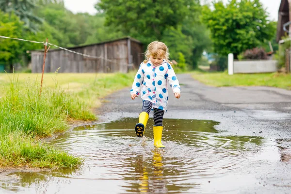 Egy kislány sárga esőcsizmában, aki esős napokon fut és sétál az ónos esőben. Aranyos boldog gyermek színes ruhák ugrás tócsa, fröccsenő vízzel, szabadtéri tevékenység — Stock Fotó