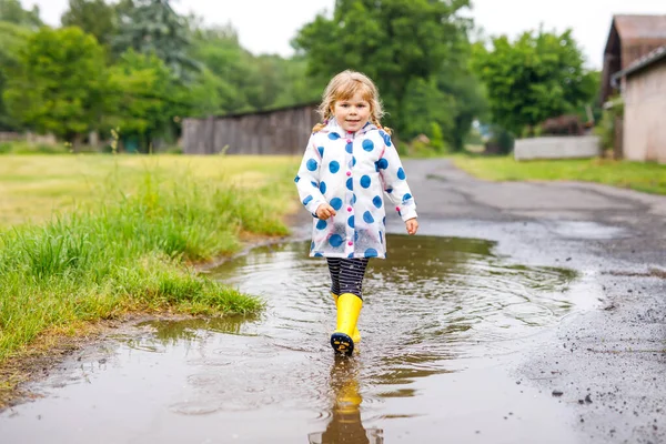 Klein peutermeisje met gele regenlaarzen, lopend en lopend tijdens de sneeuw op regenachtige bewolkte dag. Schattig vrolijk kind in kleurrijke kleren springen in plas, spetteren met water, outdoor-activiteit — Stockfoto