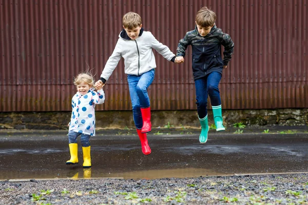 Drei Kinder, ein Kleinkind und zwei Jungen tragen rote, gelbe und grüne Regenstiefel und gehen bei Schneeregen. Glückliche Geschwister springen in eine Pfütze. Spaß im Freien, aktive Familie — Stockfoto