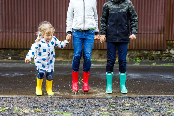 Três crianças, menina criança e dois meninos crianças vestindo botas de chuva vermelhas, amarelas e verdes e andando durante o trenó. Irmãos felizes a saltar para a poça. Divertindo-se ao ar livre, família ativa — Fotografia de Stock