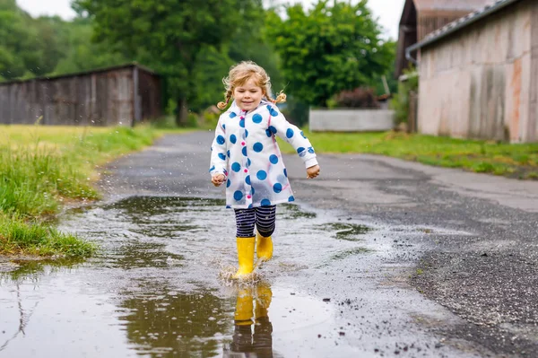 Piccola bambina che indossa stivali da pioggia gialli, correndo e camminando durante la nebbia nella giornata nuvolosa piovosa. Carino bambino felice in vestiti colorati saltare in pozzanghera, schizzi con acqua, attività all'aperto — Foto Stock