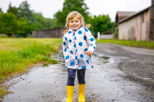 Piccola bambina che indossa stivali da pioggia gialli, correndo e camminando durante la nebbia nella giornata nuvolosa piovosa. Carino bambino felice in vestiti colorati saltare in pozzanghera, schizzi con acqua, attività all'aperto — Foto Stock