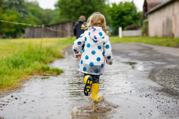 İki çocuk, küçük kız ve çocuk kırmızı ve sarı çizme giyiyorlar, yağmurluk sırasında yürüyorlar. Mutlu kardeşler, kız ve erkek kardeşler gölete atlıyor. Dışarıda eğlenmek, dışarıda aktif aile olmak. — Stok fotoğraf