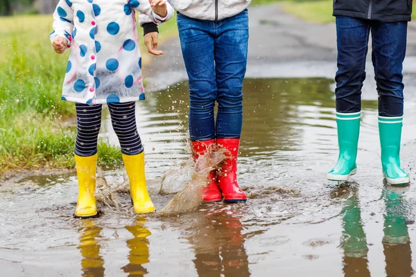 Three children, toddler girl and two kids boys wearing red, yellow and green rain boots and walking during sleet. Happy siblings jumping into puddle. Having fun outdoors, active family — Stock Photo, Image