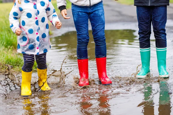 Tři děti, batole a dva kluci v červených, žlutých a zelených holínkách do deště a při chůzi během plískanice. Šťastní sourozenci skáčou do kaluže. Bavíte se venku, aktivní rodina — Stock fotografie