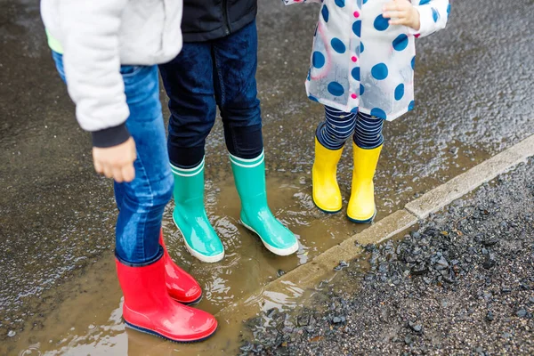 Close-up of three children, toddler girl and two kids boys wearing red, yellow and green rain boots and walking during sleet. Happy siblings jumping into puddle. Having fun outdoors, active family — Stock Photo, Image