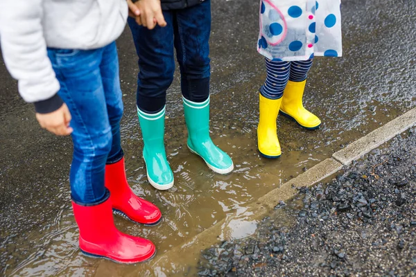 Close-up of three children, toddler girl and two kids boys wearing red, yellow and green rain boots and walking during sleet. Happy siblings jumping into puddle. Having fun outdoors, active family — Stock Photo, Image