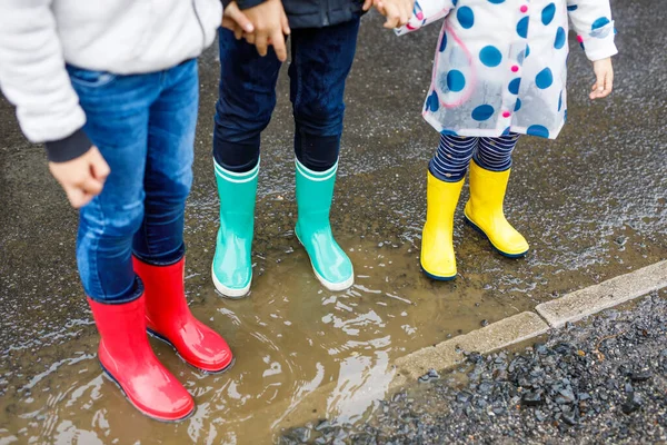 Close-up de três crianças, menina criança e dois meninos crianças vestindo botas de chuva vermelho, amarelo e verde e andando durante a manga. Irmãos felizes a saltar para a poça. Divertindo-se ao ar livre, família ativa — Fotografia de Stock