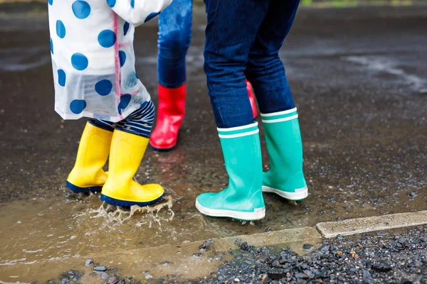 Großaufnahme von drei Kindern, einem kleinen Mädchen und zwei kleinen Jungen, die rote, gelbe und grüne Regenstiefel tragen und bei Schneeregen laufen. Glückliche Geschwister springen in eine Pfütze. Spaß im Freien, aktive Familie — Stockfoto