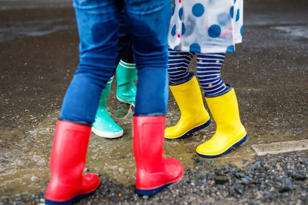 Close-up of three children, toddler girl and two kids boys wearing red, yellow and green rain boots and walking during sleet. Happy siblings jumping into puddle. Having fun outdoors, active family — Stock Photo, Image