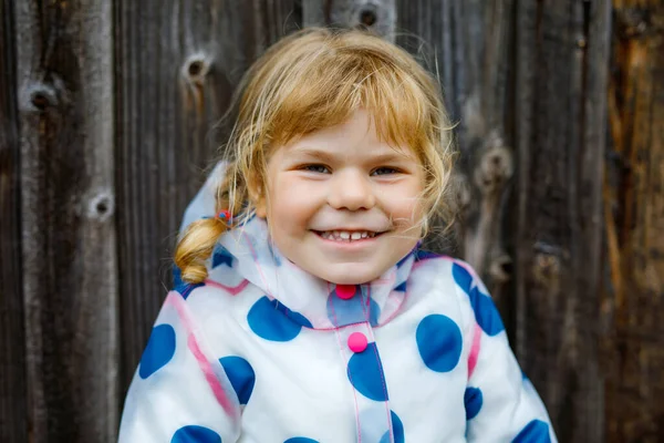 Außenporträt eines glücklich lächelnden kleinen Mädchens mit Regenjacke an einem regnerischen, bewölkten Tag. Nettes gesundes Kind in bunten Kleidern Outdoor-Aktivität — Stockfoto