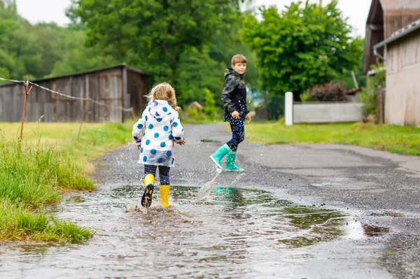 Dvě děti, batole a chlapec v červených a žlutých holínkách do deště, chodící během plískanice. Šťastní sourozenci, bratr a sestra skáčou do kaluže. Bavíte se venku, aktivní rodina venku — Stock fotografie