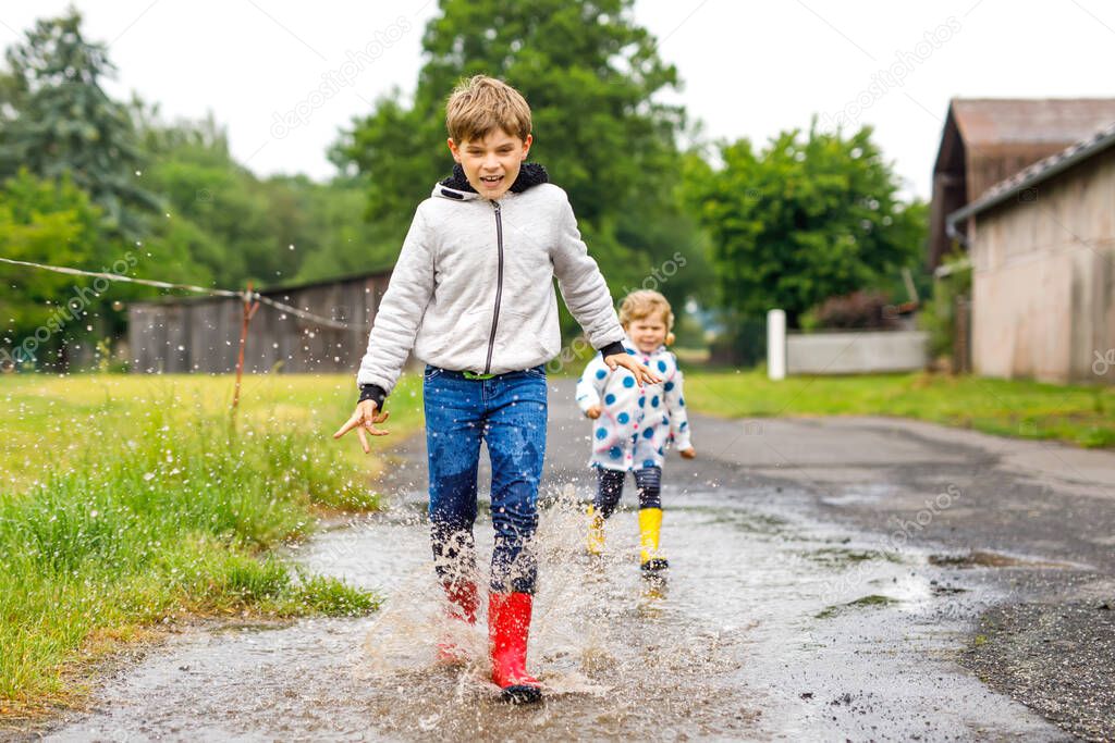 Two children, toddler girl and kid boy wearing red and yellow rain boots, walking during sleet. Happy siblings, brother and sister jumping into puddle. Having fun outdoors, active family outdoors