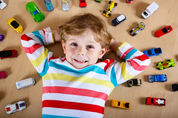 Schöne blonde Junge spielen mit vielen Spielzeugautos drinnen. Glücklicher gesunder Junge mit Spaß während der Coronavirus-Quarantäne-Pandemie. Kind allein zu Hause, geschlossene Kinderkrippe. — Stockfoto