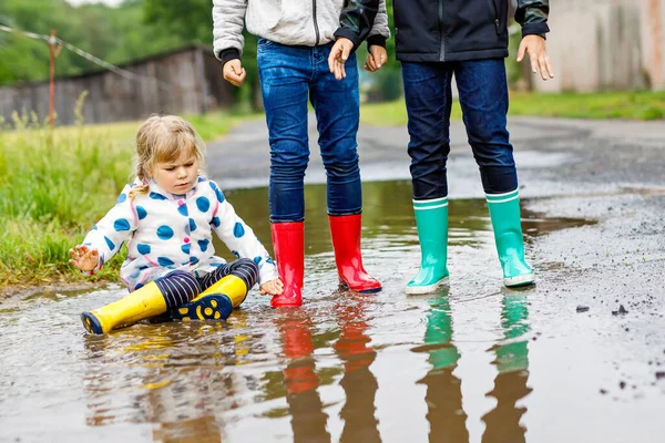 Three children, toddler girl and two kids boys wearing red, yellow and green rain boots and walking during sleet. Happy siblings jumping into puddle. Having fun outdoors, active family — Stock Photo, Image