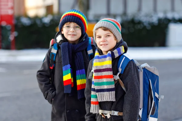 Zwei kleine Jungen der Grundschulklasse laufen bei Schneefall zur Schule. Glückliche Kinder, die Spaß haben und mit dem ersten Schnee spielen. Geschwister und Freunde mit Rucksack in bunter Winterkleidung. — Stockfoto