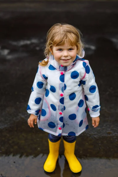 Little toddler girl wearing yellow rain boots, running and walking during sleet on rainy cloudy day. Cute happy child in colorful clothes jumping into puddle, splashing with water, outdoor activity — Stock Photo, Image