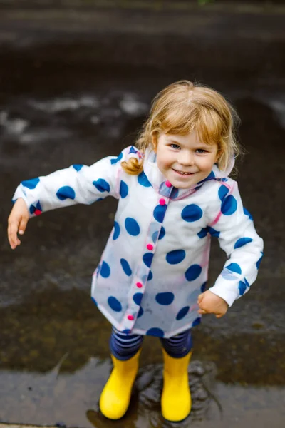 Little toddler girl wearing yellow rain boots, running and walking during sleet on rainy cloudy day. Cute happy child in colorful clothes jumping into puddle, splashing with water, outdoor activity — Stock Photo, Image
