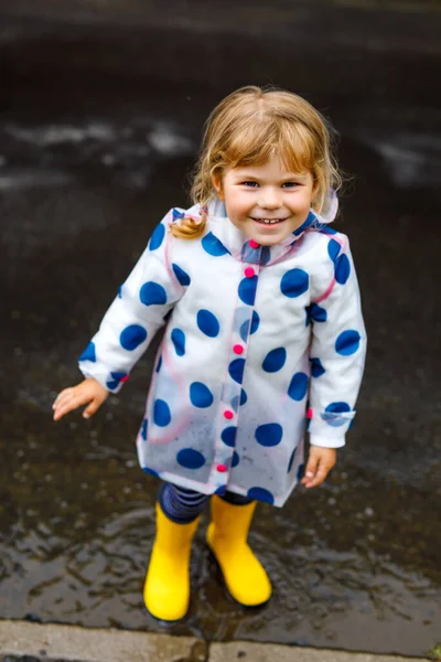 Little toddler girl wearing yellow rain boots, running and walking during sleet on rainy cloudy day. Cute happy child in colorful clothes jumping into puddle, splashing with water, outdoor activity — Stock Photo, Image