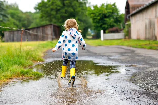 Petite fille en bas âge portant des bottes de pluie jaunes, courant et marchant pendant la neige fondante par temps de pluie nuageux. Enfant heureux mignon en vêtements colorés sautant dans la flaque d'eau, éclaboussures d'eau, activité de plein air — Photo