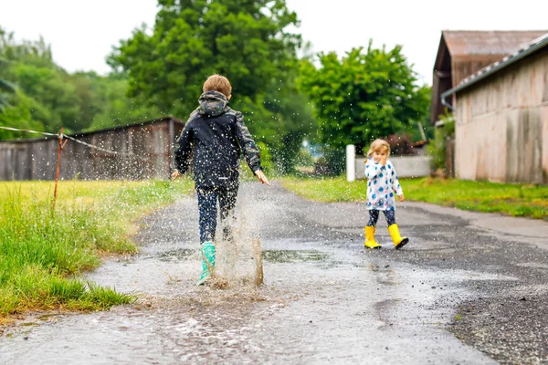 Dos niños, niña y niño con botas de lluvia rojas y amarillas, caminando durante el aguanieve. Felices hermanos, hermano y hermana saltando al charco. Divertirse al aire libre, familia activa al aire libre —  Fotos de Stock