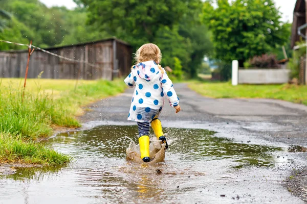 Malá batolata ve žlutých holínkách do deště, běhající a chodící během deštivého oblačného dne. Roztomilé šťastné dítě v barevných šatech skákání do kaluže, cákání vodou, outdoorové aktivity — Stock fotografie