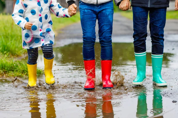 Três crianças, menina criança e dois meninos crianças vestindo botas de chuva vermelhas, amarelas e verdes e andando durante o trenó. Irmãos felizes a saltar para a poça. Divertindo-se ao ar livre, família ativa — Fotografia de Stock