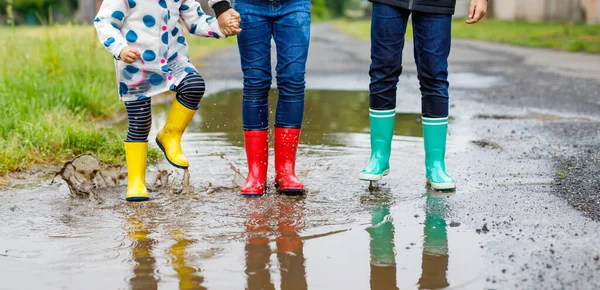 Three children, toddler girl and two kids boys wearing red, yellow and green rain boots and walking during sleet. Happy siblings jumping into puddle. Having fun outdoors, active family — Stock Photo, Image