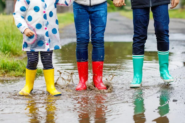 Üç çocuk, yeni yürümeye başlayan bir kız ve iki çocuk kırmızı, sarı ve yeşil çizme giyiyorlar ve yağmurluk sırasında yürüyorlar. Mutlu kardeşler gölete atlıyor. Dışarıda eğleniyor, aktif bir aile. — Stok fotoğraf