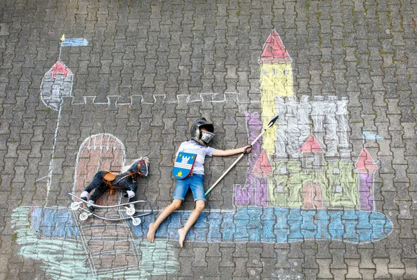 Malý aktivní chlapec kreslí rytířský hrad a pevnost s barevnými křídami na asfaltu. Šťastné dítě v helmě a s oštěpem baví s hraním rytířské hry a malování — Stock fotografie