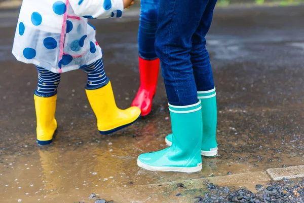 Großaufnahme von drei Kindern, einem kleinen Mädchen und zwei kleinen Jungen, die rote, gelbe und grüne Regenstiefel tragen und bei Schneeregen laufen. Glückliche Geschwister springen in eine Pfütze. Spaß im Freien, aktive Familie — Stockfoto