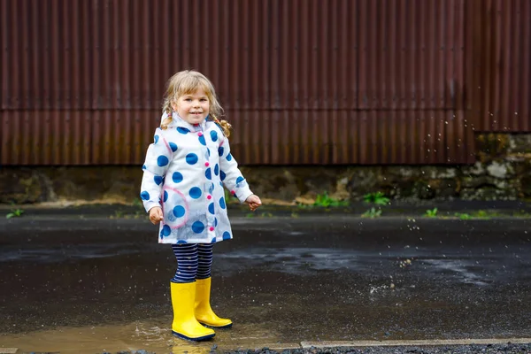 Μικρό κοριτσάκι φορώντας κίτρινες μπότες βροχής, τρέχει και περπατά κατά τη διάρκεια sleet στις βροχερές συννεφιασμένες ημέρες. Χαριτωμένο χαρούμενο παιδί σε πολύχρωμα ρούχα άλμα σε λακκούβα, πιτσιλίσματα με νερό, υπαίθρια δραστηριότητα — Φωτογραφία Αρχείου