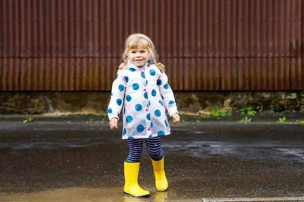 Dziewczynka w żółtych butach, biegająca i spacerująca w deszczowy pochmurny dzień. Cute szczęśliwy dziecko w kolorowe ubrania skoków do kałuży, pluskanie wodą, aktywność na świeżym powietrzu — Zdjęcie stockowe