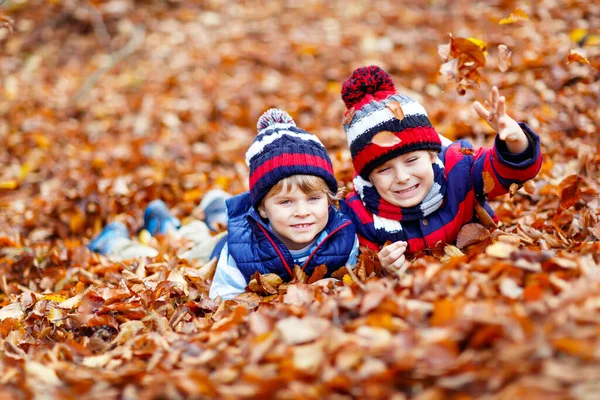 两个孪生兄弟穿着五彩斑斓的衣服躺在秋天的树叶上.快乐的兄弟姐妹孩子们在秋天的森林或公园里玩的开心.戴着休闲装的帽子和围巾。朋友们一起玩. — 图库照片