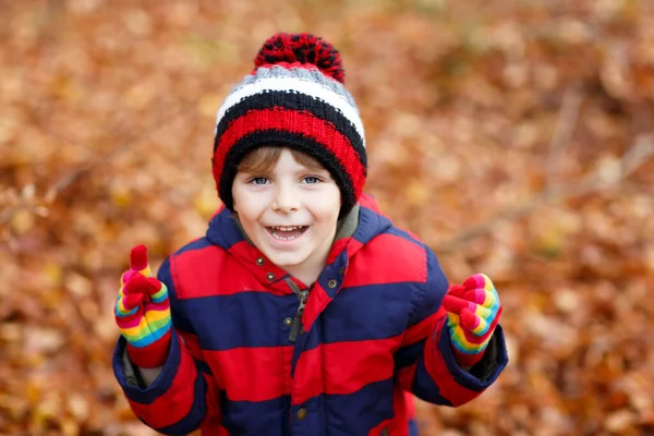Portrét šťastného roztomilého chlapečka s podzimními listy na pozadí v barevném oblečení. Vtipné dítě baví v podzimním lese nebo parku. Usmívající se dítě v módní a barevné rukavice. — Stock fotografie