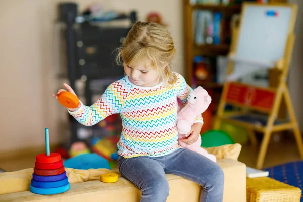 Мила маленька дівчинка-малюк грає сама з барвистою дерев'яною веселкою пірамідою та іграшками вдома або в дитячій кімнаті. Щаслива здорова дитина розважається в дитячому садку або дошкільному догляді — стокове фото