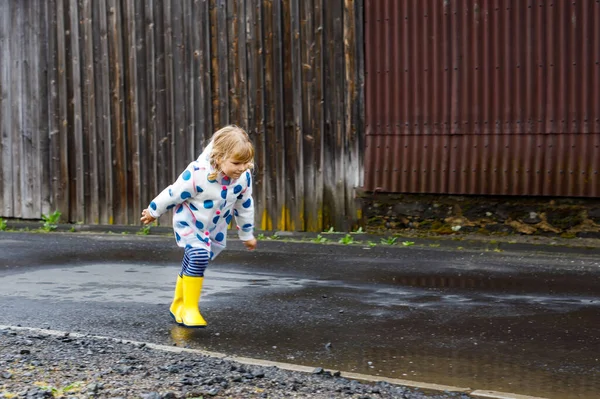 Niña pequeña con botas de lluvia amarillas, corriendo y caminando durante el aguanieve en el día nublado lluvioso. Lindo niño feliz en ropa colorida saltando en el charco, salpicaduras de agua, actividad al aire libre —  Fotos de Stock