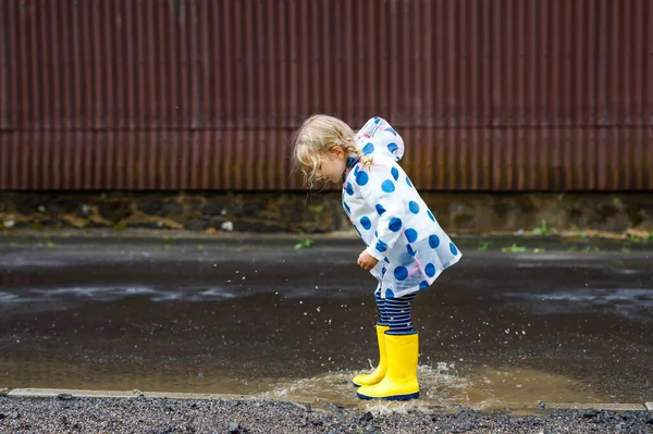 Menina pequena usando botas de chuva amarelas, correndo e andando durante o trenó em dia nublado chuvoso. Criança feliz bonito em roupas coloridas pulando em poça, salpicando com água, atividade ao ar livre — Fotografia de Stock