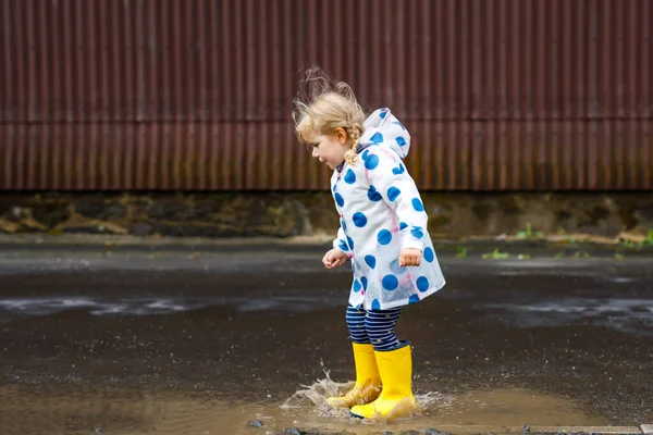 Klein peutermeisje met gele regenlaarzen, lopend en lopend tijdens de sneeuw op regenachtige bewolkte dag. Schattig vrolijk kind in kleurrijke kleren springen in plas, spetteren met water, outdoor-activiteit — Stockfoto