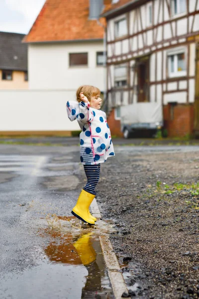 Little toddler girl wearing yellow rain boots, running and walking during sleet on rainy cloudy day. Cute happy child in colorful clothes jumping into puddle, splashing with water, outdoor activity — Stock Photo, Image