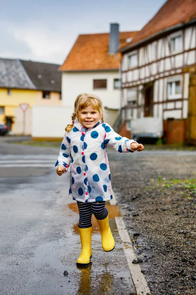 Liten småbarn flicka bär gula regnstövlar, springa och gå under slask på regnig molnig dag. Söt glad barn i färgglada kläder hoppar i pöl, stänk med vatten, utomhus aktivitet — Stockfoto