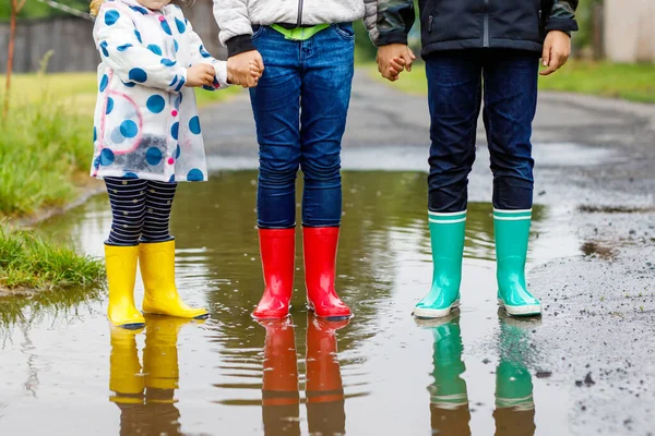 Üç çocuk, yeni yürümeye başlayan bir kız ve iki çocuk kırmızı, sarı ve yeşil çizme giyiyorlar ve yağmurluk sırasında yürüyorlar. Mutlu kardeşler gölete atlıyor. Dışarıda eğleniyor, aktif bir aile. — Stok fotoğraf