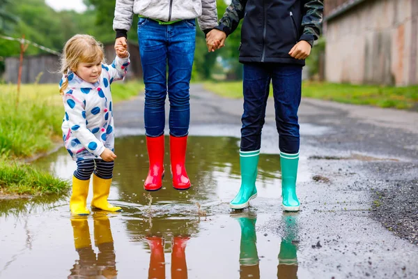 Tres niños, una niña pequeña y dos niños con botas de lluvia rojas, amarillas y verdes y caminando durante el aguanieve. Felices hermanos saltando al charco. Divertirse al aire libre, familia activa —  Fotos de Stock