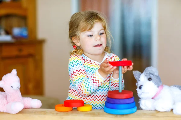 Cute little toddler girl playing alone with colorful wooden rainbow pyramid and toys at home or nursery. Happy healthy child having fun in kindergarten or preschool daycare — Stock Photo, Image