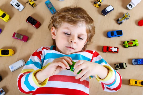 Härligt blond pojke leker med massor av leksaksbilar inomhus. Glad frisk pojke som har roligt under en pandemisk karantänsjukdom. Ensamstående barn hemma, stängd barnkammare. — Stockfoto