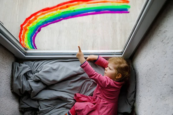 Adoralbe kleines Mädchen mit Regenbogen bemalt mit bunten Fensterfarben während der Coronavirus-Quarantäne-Pandemie. Kind bemalt Regenbogen und Herzen auf der ganzen Welt mit Worten. — Stockfoto