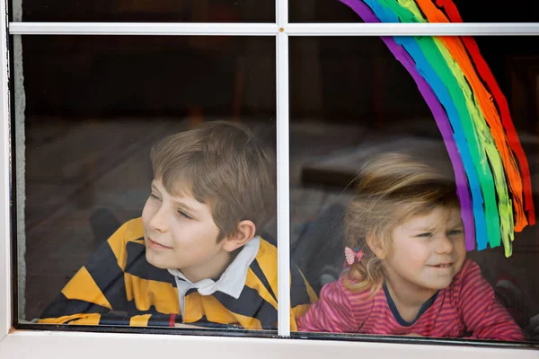 Två barn, skolpojke och småbarn flicka med regnbåge målade med färgglada fönster färg under pandemisk coronavirus karantän. Barn måla regnbågar med orden Låt alla må bra — Stockfoto
