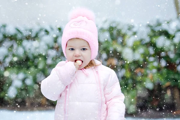 Portret van een klein peutermeisje dat 's winters buiten loopt. Leuke peuter die zoete lolly snoep eet. Kind heeft plezier op koude sneeuwdag. Het dragen van warme baby roze kleren en hoed met bobbels. — Stockfoto